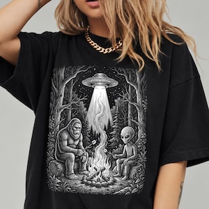 May include: A black t-shirt featuring a detailed graphic print of a Bigfoot and an alien sitting by a campfire in a forest. Above them, a UFO hovers in the night sky, casting a beam of light down toward the fire. Both figures hold sticks.