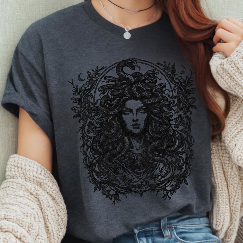 Op de afbeelding: Donkergrijs t-shirt met een gedetailleerde zwarte illustratie van Medusa. De afbeelding toont Medusa's gezicht, omringd door slangen en bloemmotieven. Het t-shirt heeft korte mouwen en een ronde hals.