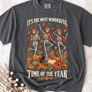 May include: A charcoal gray t-shirt featuring a graphic of three skeletons wearing orange beanies and scarves, dancing among pumpkins, autumn leaves, and a black cat. The text reads, "IT'S THE MOST WONDERFUL TIME OF THE YEAR" in a cream-colored font.