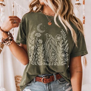 Pode incluir: Camiseta verde-oliva com um desenho branco de um coelho olhando para uma lua crescente, rodeado por elementos florais. A camiseta tem mangas curtas e um corte folgado. A modelo está usando jeans azuis e um cinto marrom.
