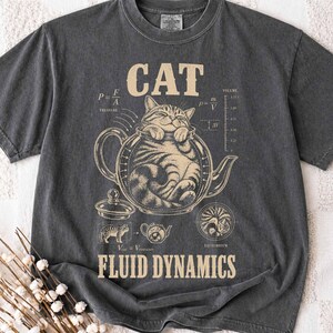 Puede incluir: Una camiseta gris carbón con un gráfico beige de un gato dentro de una tetera. El diseño incluye el texto CAT y FLUID DYNAMICS, junto con ecuaciones físicas, una escala de volumen e ilustraciones pequeñas de un gato y una tetera.