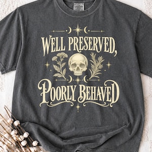 May include: A charcoal grey short-sleeve t-shirt featuring a cream-colored graphic print. The design includes a central human skull flanked by botanical illustrations, stars, and crescent moons, with the text Well Preserved, Poorly Behaved.