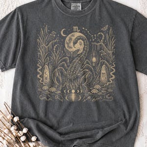 Könnte beinhalten: Ein anthrazitfarbenes T-Shirt mit einem beigen Grafikdruck eines großen Skorpions unter einem Mond. Das Design zeigt einen Nachtfalter, eine Laterne, zwei Steine mit Tierkreissymbolen, Schilfrohr, Seerosen und eine Spiegelung im Wasser.