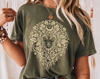 May include: An olive green short-sleeve t-shirt featuring a large, circular cream-colored graphic print on the chest. The design depicts a stylized face surrounded by intricate oak leaves, acorns, and a crescent moon shape at the top center.