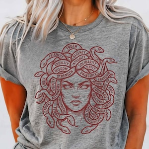 以下が含まれることがあります： ヘザーグレーのTシャツで、髪が蛇になっている顔のダークレッドのグラフィックが描かれています。蛇には、Truth、Decency、Integrity、Solidarity、Unity、Principles、Freedom、Honor、Courage、Resilience、Power、Humanity、Love、Visibility、Support、Female、Independence、Autonomy、Dignity、Fight、Wisdom、Strength、Equality、Fairnessといった言葉が組み込まれています。