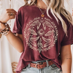 May include: Burgundy-colored t-shirt featuring a circular design of stylized birds and celestial elements. The graphic is in a cream color, creating a contrast against the shirt's deep red hue. The shirt has a relaxed fit and short sleeves.