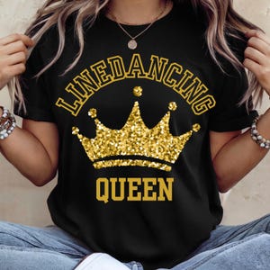 以下が含まれることがあります： 金色のグリッタークラウンのデザインと「LINEDANCING QUEEN」の文字が金色でプリントされた黒いTシャツ。デザインは前面の中央に配置されています。Tシャツはクルーネックです。