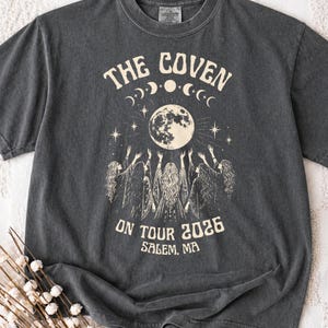 May include: A charcoal grey short-sleeve t-shirt featuring a cream-coloured graphic of a full moon, stars, and moon phases above three figures with raised arms. The text reads THE COVEN ON TOUR 2026 SALEM, MA in a stylised serif font.