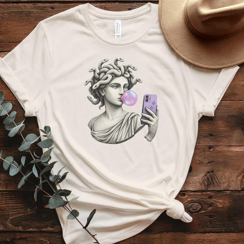 May include: Off-white t-shirt with a graphic of Medusa, styled as a classical statue, blowing a bubblegum bubble and taking a selfie with a purple phone. The shirt has a round neck and short sleeves.