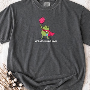 May include: A charcoal grey short-sleeve t-shirt featuring a graphic of a green frog wearing a red cape, holding a pink feather duster and a blue spray bottle. Below the frog, the text reads WE RAGE CLEAN AT DAWN in white capital letters.