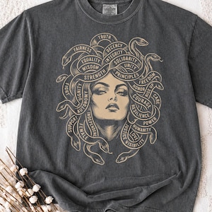 May include: A charcoal grey t-shirt featuring a beige graphic of a face with snakes for hair. The snakes are intertwined with words including Truth, Fairness, Equality, Wisdom, Strength, Dignity, Autonomy, Independence, Female, Support, Fight, Solidarity, Integrity, Unity, Principles, Honor, Freedom, Rise Up, Care, Courage, Resilience, Power, Humanity, Love, and Visibility.