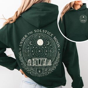 May include: Dark green hoodie with a white graphic on the back featuring Stonehenge, a full moon, stars, and the text "UNDER THE SOLSTICE MOON." A smaller version of the design is on the front.