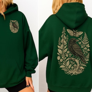 May include: A dark forest green hooded sweatshirt featuring a graphic print of a black crow perched on a branch, surrounded by botanical elements including a moth, ferns, mushrooms, and flowers, displayed on both the front and back.
