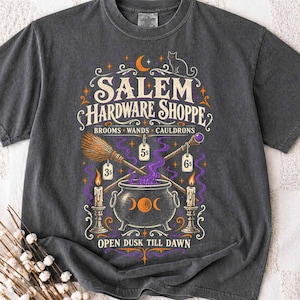 May include: A charcoal gray t-shirt featuring a graphic print with the text SALEM HARDWARE SHOPPE, BROOMS, WANDS, CAULDRONS, and OPEN DUSK TILL DAWN. The design includes a cauldron, a broom, a wand, candles, a cat, and price tags labeled 3s, 5s, and 6s.