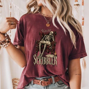 Pode incluir: Uma t-shirt de manga curta cor de vinho com um gráfico de um esqueleto sentado numa poltrona vermelha a ler um livro, com o texto "SLEEPY" acima e "SCRIBBLER" abaixo, incluindo uma pequena vela acesa e papéis soltos no chão.