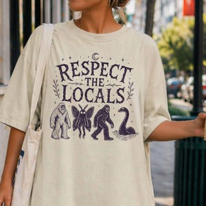 Puede incluir: Una camiseta de manga corta color crema con un estampado gráfico en color morado oscuro. El texto "RESPECT THE LOCALS" aparece sobre ilustraciones de un yeti, un mothman, un sasquatch y el monstruo del lago Ness, con luna y estrellas.