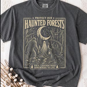 May include: A charcoal grey t-shirt featuring a cream-coloured graphic print with the text "PROTECT OUR HAUNTED FORESTS" and "FOLK LEGENDS NEED SOMEWHERE TO LIVE." The illustration depicts a crescent moon, a moth, a ghost, a cryptid, and a figure.