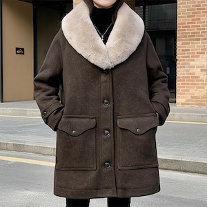 Puede incluir: Un abrigo de lana marrón con cuello de piel sintética beige claro. El abrigo tiene dos bolsillos delanteros grandes, cierre de botones y un corte a media pierna. El cuello contrasta con el marrón más oscuro del abrigo.