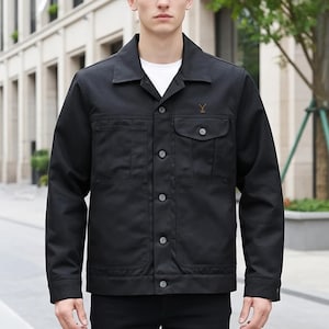 Puede incluir: Chaqueta negra con botones, dos bolsillos en el pecho y dos bolsillos inferiores. La chaqueta tiene cuello y un pequeño diseño bordado en el pecho. El modelo lleva una camiseta blanca debajo. La chaqueta es de estilo casual.