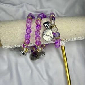 May include: Three beaded bracelets with purple and clear beads, gold accents, and silver charms. One charm reads "IN MEMORIAM," and another reads "PAMELA VALIEVA." The bracelets are displayed on a cream-colored surface.