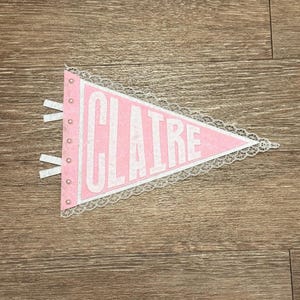 May include: Pink pennant banner with the name "CLAIRE" in white block letters. The banner has a white lace trim and small pearl accents. Two white ribbons are attached to the left side. The banner is on a wooden surface.