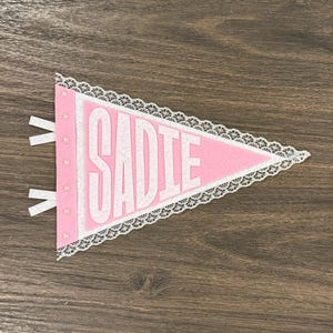May include: A triangular pink felt pennant featuring the name SADIE in white block letters. The pennant is bordered by white lace trim and includes a vertical strip on the left side with small white beads and two white fabric hanging loops.