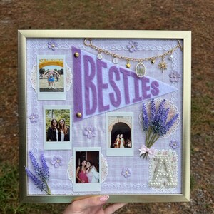 Puede incluir: Un tablero de recuerdos enmarcado con un marco dorado, con una bander&iacute;n de fieltro morado que dice "BESTIES". El tablero incluye varias fotos Polaroid, ramitas de lavanda y adornos decorativos. El fondo es de tela morada clara.