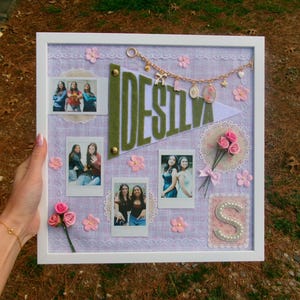 Puede incluir: Un tablero de recuerdos enmarcado en blanco con fotos, flores rosas y el nombre "DESILVA" en letras de fieltro verde. El tablero est&aacute; decorado con encaje, una bandera y una cadena dorada con dijes.