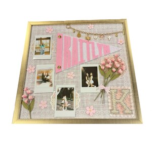 Puede incluir: Un tablero de recuerdos enmarcado con un marco dorado, con un bander&iacute;n rosa que dice "RAITTLYN". El tablero incluye varias fotos peque&ntilde;as, flores rosas y una "K" decorativa. Una cadena de amuletos cuelga sobre el bander&iacute;n.