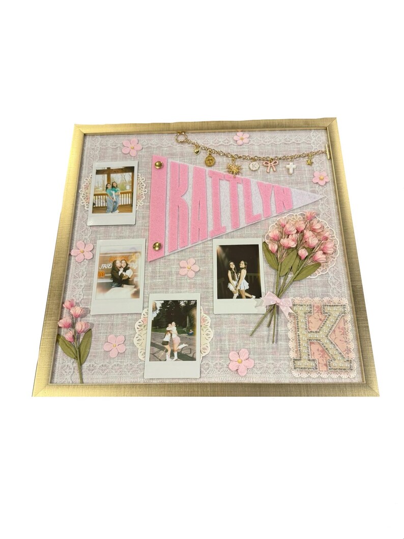 Puede incluir: Un tablero de recuerdos enmarcado con un marco dorado, con un bander&iacute;n rosa que dice "RAITTLYN". El tablero incluye varias fotos peque&ntilde;as, flores rosas y una "K" decorativa. Una cadena de amuletos cuelga sobre el bander&iacute;n.