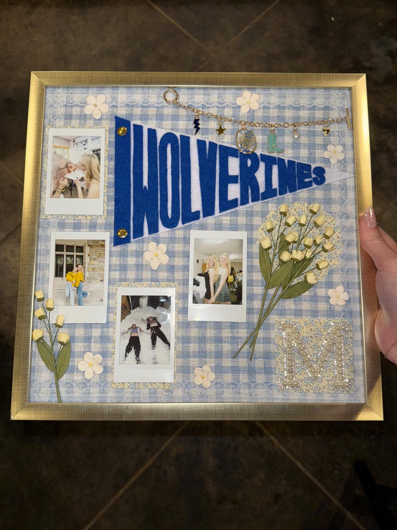 Puede incluir: Un collage enmarcado con un fondo de cuadros vichy azul y blanco. Presenta cuatro fotos polaroid, un bander&iacute;n azul con "Wolverines" en blanco y flores y adornos decorativos. El marco es dorado.