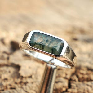 Könnte beinhalten: Ein Silberring mit einem rechteckigen Moosachatstein. Der Stein hat grüne und braune Einschlüsse, gefasst in einem polierten Silberband. Der Ring wird auf einer Holzoberfläche präsentiert.