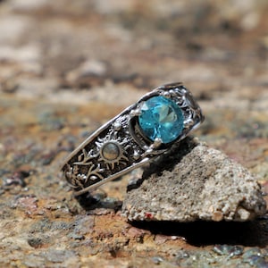 May include: A silver-toned ring featuring a round, faceted bright blue gemstone set in the center. The band is decorated with embossed celestial motifs, including a sun and star-like patterns, with a dark, oxidized finish in the recessed areas.