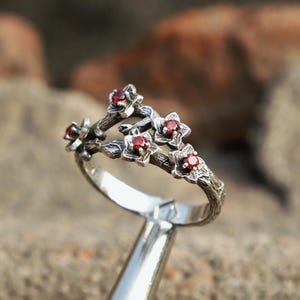 Könnte beinhalten: Ein silberner Ring mit floralem Design und kleinen, roten Edelsteinen. Das Ringband hat ein strukturiertes, astähnliches Aussehen, und die Blüten sind oben angeordnet. Die Edelsteine sind tiefrot und setzen einen Farbakzent auf dem Silber.