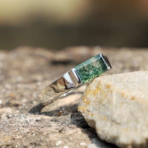 May include: A silver ring with a rectangular, moss-green stone. The ring is set on a rough, textured stone surface, with a blurred background. The ring's band is polished and reflects light.