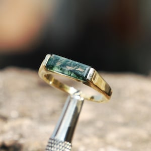 Op de afbeelding: Een gouden ring met een rechthoekige groene steen. De ring heeft een eenvoudige band en een bezel setting. De steen heeft een uniek patroon van groene en zwarte insluitsels. De ring wordt vastgehouden door een zilveren gereedschap.
