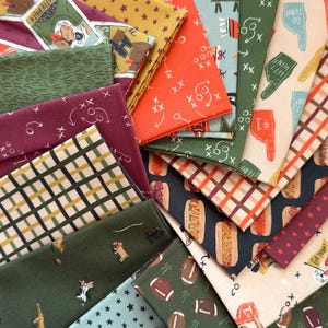 May include: A collection of folded fabric swatches featuring various sports-themed patterns including football plays, brown footballs, hot dogs, foam fingers, and dogs, alongside geometric star and plaid prints in shades of orange, green, and blue.