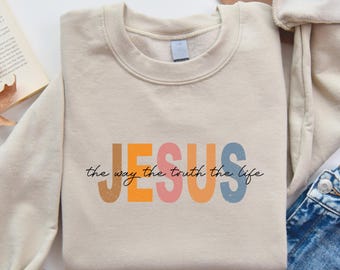 May include: A sand-colored sweatshirt with the word "JESUS" in colorful block letters. Below the word "JESUS" is the phrase "the way the truth the life" in a handwritten font. The sweatshirt is folded with the sleeves rolled up.