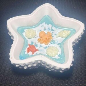 Puede incluir: Un plato blanco en forma de estrella con un borde texturizado. El interior presenta una resina azul claro con adornos de criaturas marinas, incluyendo un pulpo naranja, una tortuga roja y conchas blancas.