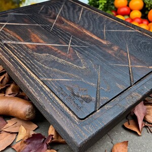 Puede incluir: Tabla de cortar de madera teñida oscura con forma rectangular y diseño geométrico tallado. La tabla tiene una ranura para jugos empotrada y las palabras "True Wood" están grabadas en la esquina. Rodeada de hojas de otoño.