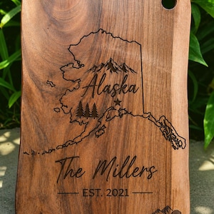May include: A rectangular brown wooden cutting board with a live edge, featuring a laser-engraved map of Alaska, mountain and tree icons, a star, the text Alaska, The Millers, EST. 2021, and a small mountain logo with the text True Wood.