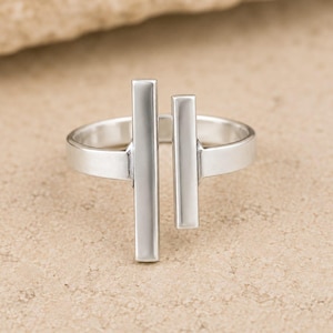 Peut inclure: Une bague argentée dotée d'un anneau ouvert avec deux barres rectangulaires verticales de longueurs différentes positionnées parallèlement au centre. Le métal présente une finition lisse et polie qui reflète la lumière sur toute sa surface.