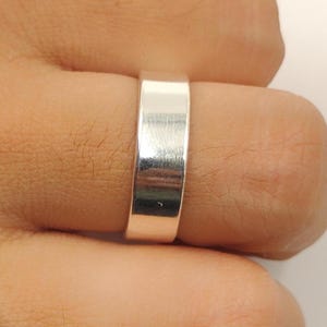 Peut inclure: Gros plan d'une bague argentée sur un doigt. La bague est une simple bande avec une surface plate. La bague est en métal brillant et réfléchissant.