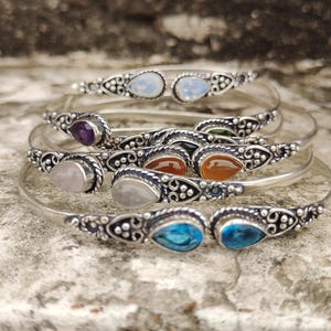 Puede incluir: Una colección de brazaletes plateados, cada uno adornado con una gema diferente. Las gemas incluyen amatista, piedra lunar, cornalina y topacio azul, engastadas en engastes decorativos de estilo filigrana. Los brazaletes se exhiben sobre una superficie gris texturizada.