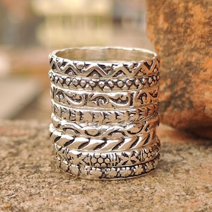 May include: A stack of seven silver rings, each with a different embossed pattern. The designs include geometric shapes, floral patterns, and textured lines. The rings are stacked, creating a layered effect. The rings are made of silver.