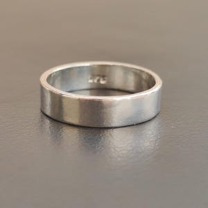 Op de afbeelding: Een gepolijste zilverkleurige bandring met een plat, glad oppervlak. De binnenkant van de ring is voorzien van een duidelijke gravure van het getal 925, wat de zuiverheid van het metaal aangeeft. De ring heeft een eenvoudige, uniforme cirkelvorm met een reflecterende afwerking.