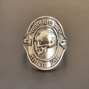 Puede incluir: Un anillo de metal plateado con una cara ovalada que presenta un relieve de calavera en el centro. El texto "memento mori" está grabado en el borde superior y "amor fati" en el borde inferior, junto a pequeños símbolos de reloj de arena y llama.