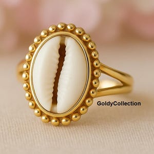 May include: A gold ring with a white cowrie shell as its focal point. The shell is oval and framed by a beaded gold border. The ring's band splits into two at the top. The text "GoldyCollection" is visible. The ring is a unique piece of jewellery.