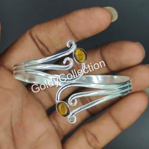 May include: Silver arm cuff with two amber gemstones. The cuff has an open design with decorative swirls and the text "Goldy Collection". The gemstones are round and set in the silver metal.