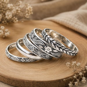 May include: A set of five silver-toned metal rings arranged in a row. Each ring features a distinct embossed pattern, including floral motifs, leaf-like textures, and geometric shapes. The rings have a polished finish with darkened recessed areas.
